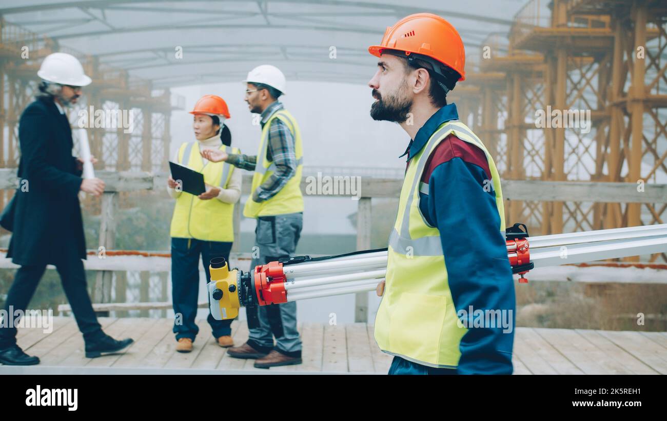 Ernsthafter männlicher Vermesser bei gleichmäßigem Gehen auf der Baustelle, der Theodolit auf die Arbeit konzentriert hält. Menschen- und Berufskonzept. Stockfoto