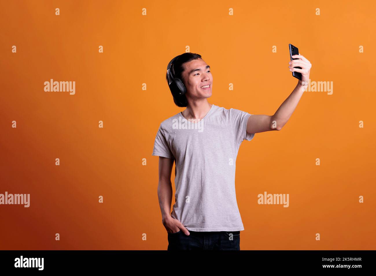 Lächelnder asiatischer Teenager in kabellosen Kopfhörern, der mit der Smartphone-Frontkamera Selfie macht. Junger, fröhlicher, attraktiver Mann in Kopfhörern, der Mobiltelefon hält und für Fotos posiert Stockfoto