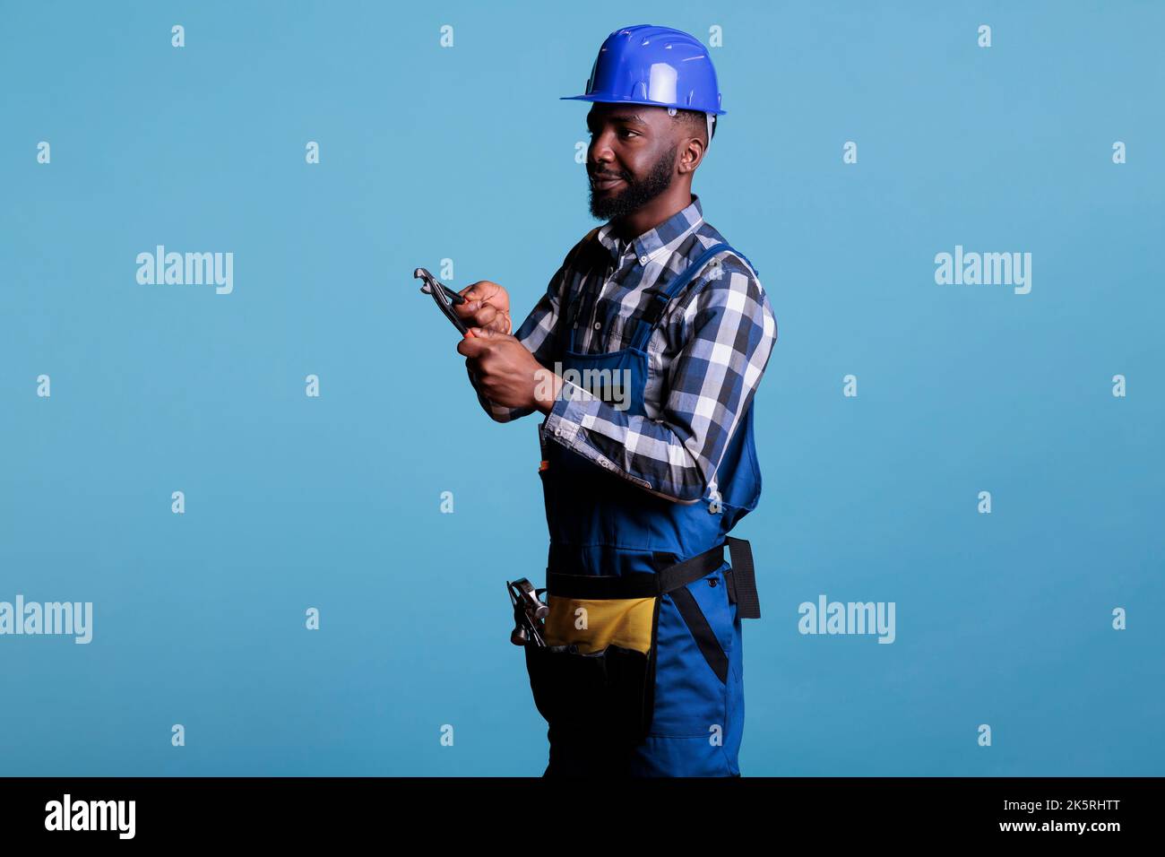 Freundliche, fröhlich lächelnde, professionelle Bauarbeiter mit Hut und Arbeitswerkzeugen auf blauem Hintergrund isoliert. afroamerikanischer Baumeister schaut zur Seite und hält Zangen. Stockfoto