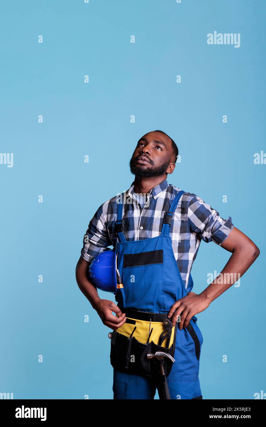 Nachdenklicher Bauarbeiter, der in einem Studiofoto vor blauem Hintergrund einen Harthut mit Overalls hält und nach oben blickt. Der Baumeister denkt über zukünftige Entscheidungen nach, die eine kurze Pause von der Arbeit machen. Stockfoto