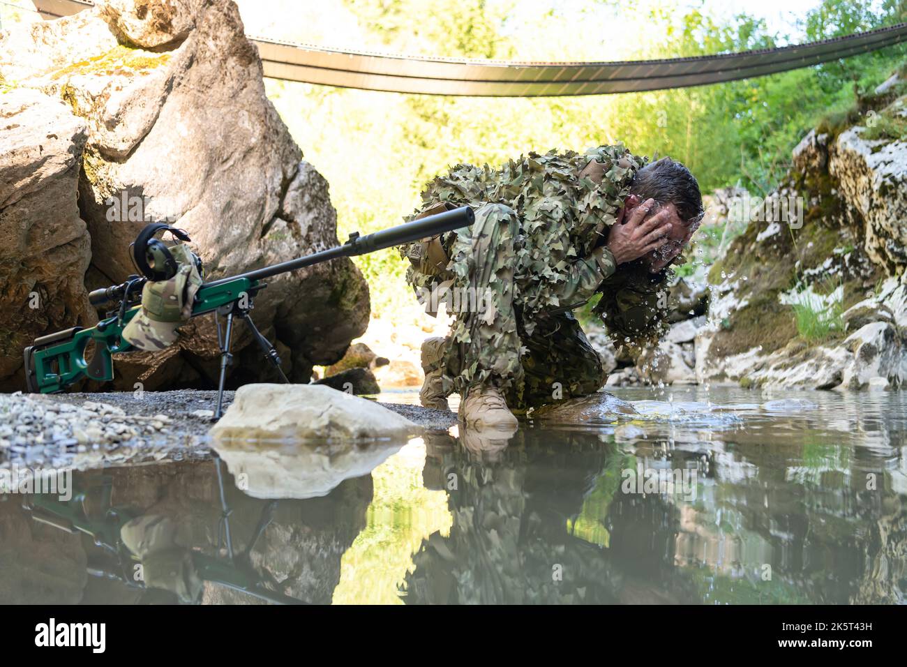 Soldat im Tarnanzug Uniform trinkt frisches Wasser aus dem Fluss. Militär-Scharfschützengewehr auf der Seite. Stockfoto