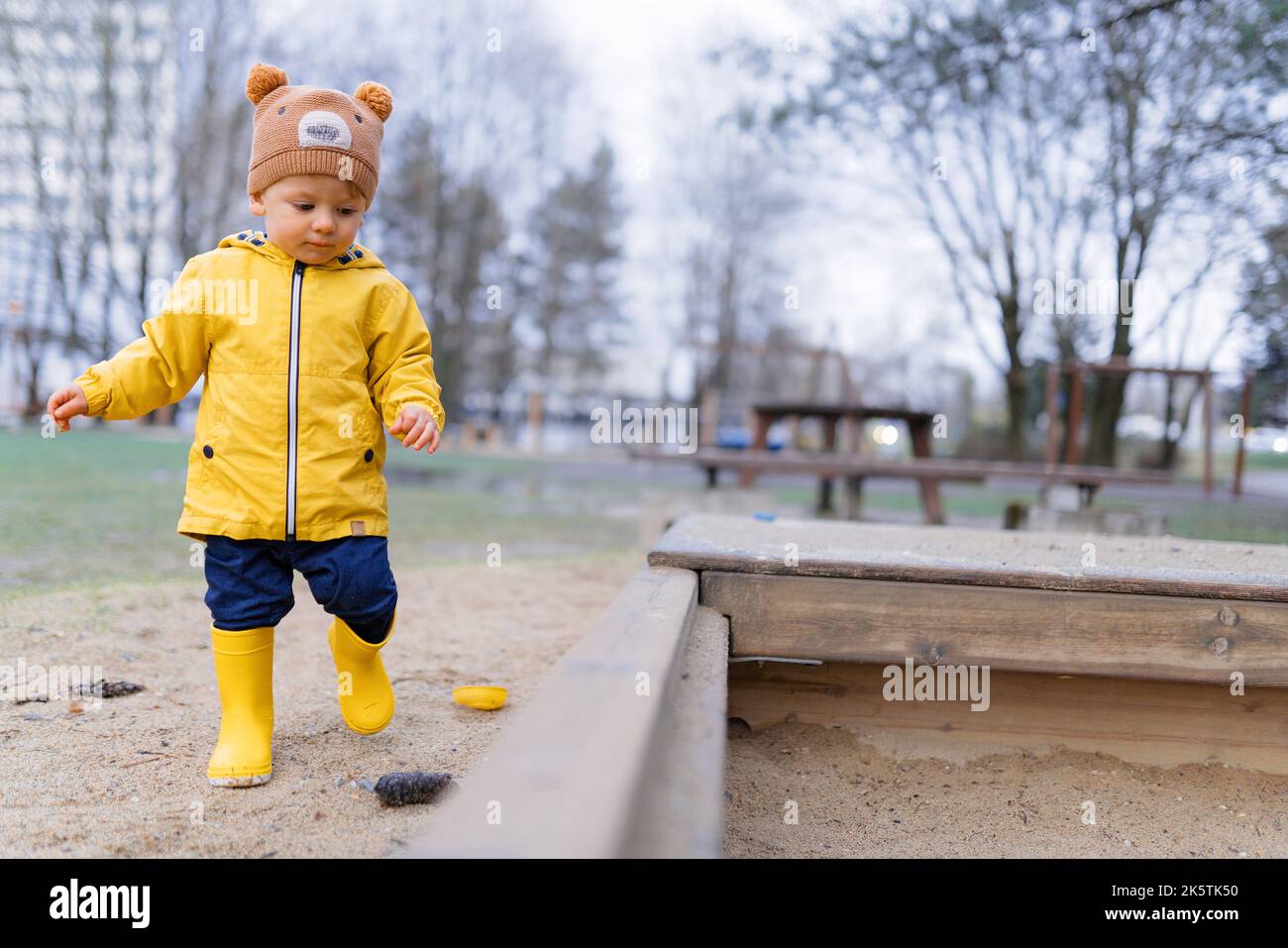 Fröhlicher kleiner Junge mit Teddybärmütze, der am Herbsttag auf dem Spielplatz im Sandkasten spielt. Stockfoto