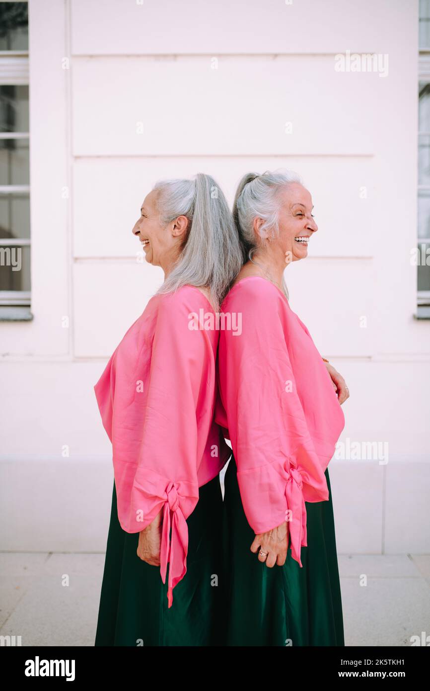 Seitenansicht von älteren Frauen, Zwillingen, in derselben Kleidung, die Rücken an Rücken steht, im Freien in der Stadt. Stockfoto