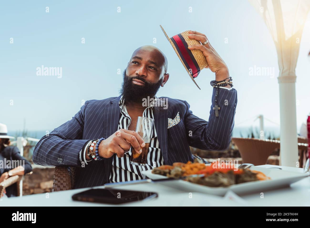 Porträt eines stämchenhaften, kahlen Afrikaners mit schwarzem Bart und in einem maßgeschneiderten Anzug, der in einem Küstenstraßenrestaurant mit einem Glas b zu Mittag gegessen hat Stockfoto