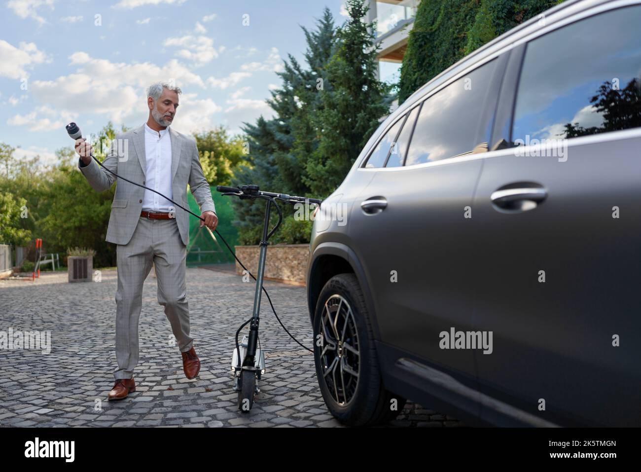 Geschäftsmann im Anzug auf dem Weg, mit Elektroscooter zu arbeiten und sein Elektroauto aufzuladen. Konzept von ökologischem Pendeln und umweltfreundlicheren Verkehrsmitteln. Stockfoto