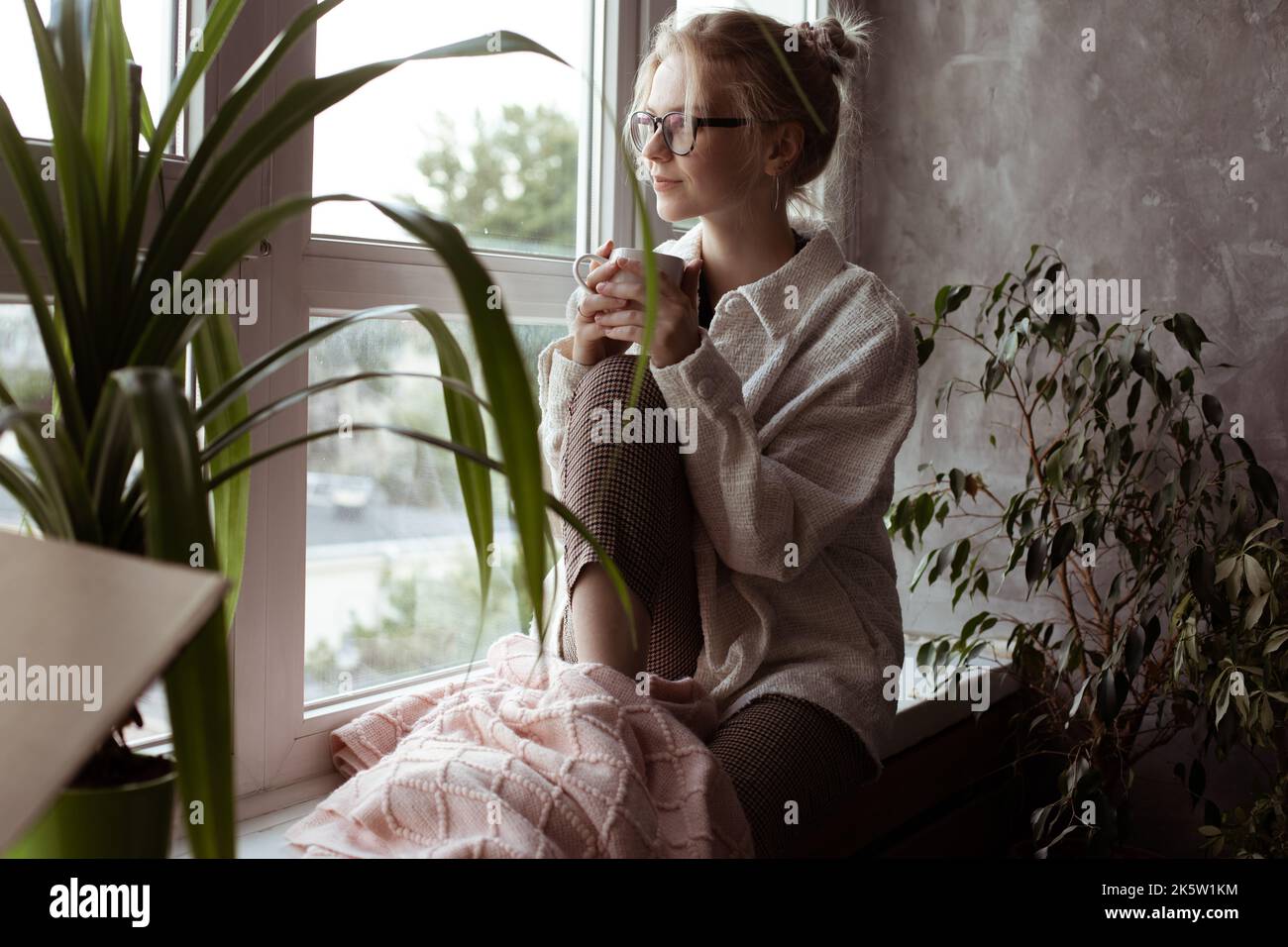Eine glückselige, ruhige, nachdenkliche junge blonde Frau mit Brille blickt mit heißem Tee aus dem Fenster. Umzug, neues Leben und Umzug Stockfoto