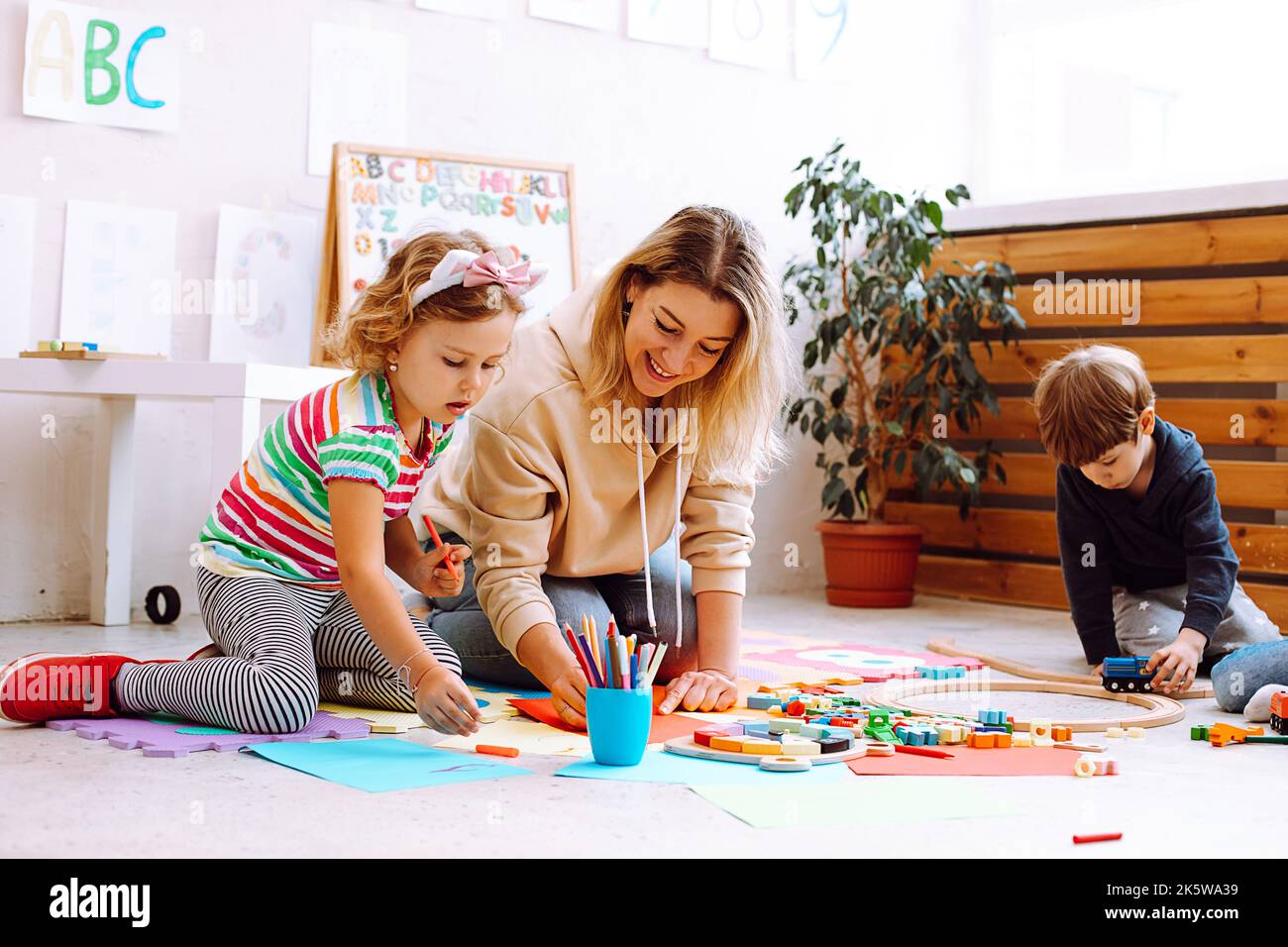 Lächelnde weibliche Psychologin spielen und testen Kinder, machen Kunsthandwerk, handgemachtes Spielzeug, Kunst im Entwicklungszentrum. Soziale Probleme Stockfoto