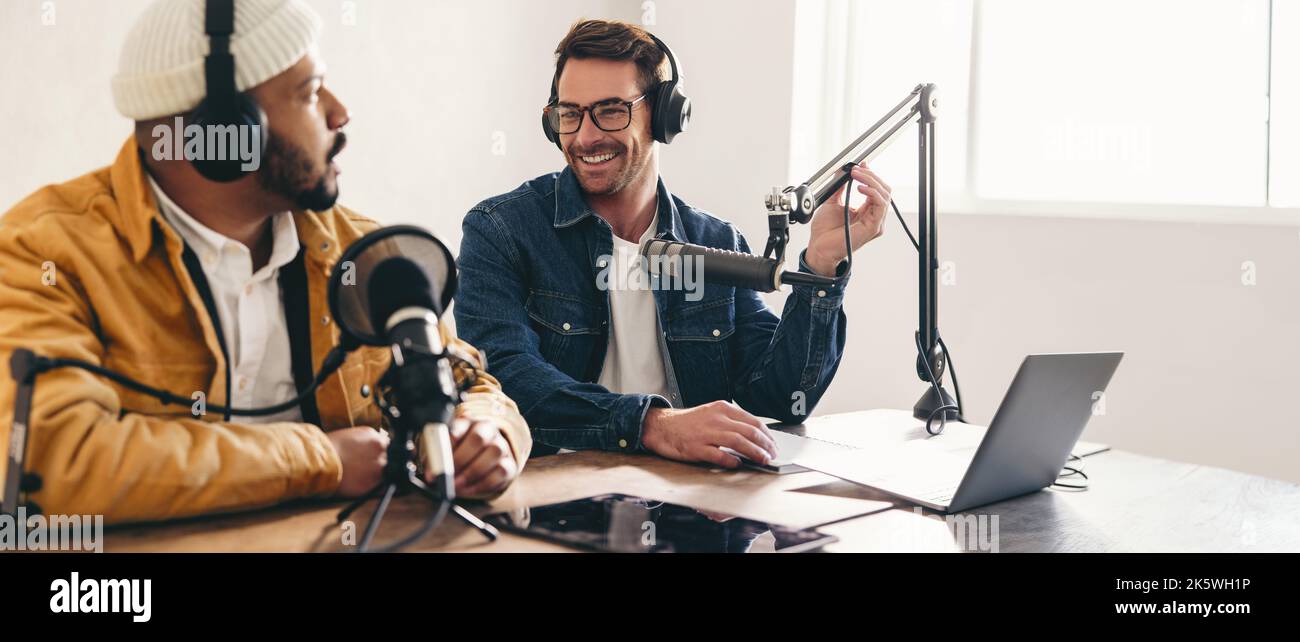 Fröhlicher Radiomoderator, der lächelt, während er einen Gast auf einem Podcast interviewst. Fröhlicher junger Mann, der zusammen mit einem Gast eine Audioübertragung moderiert. Zwei Inhaltscrea Stockfoto