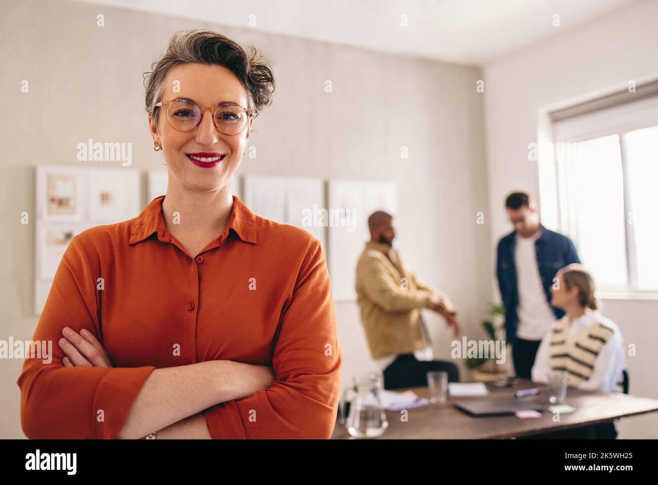 Porträt einer reifen Geschäftsfrau, die lächelt an der Kamera, während sie mit gekreuzten Armen in einem Büro steht. Glückliche Geschäftsfrau arbeitet in einer kreativen Stockfoto