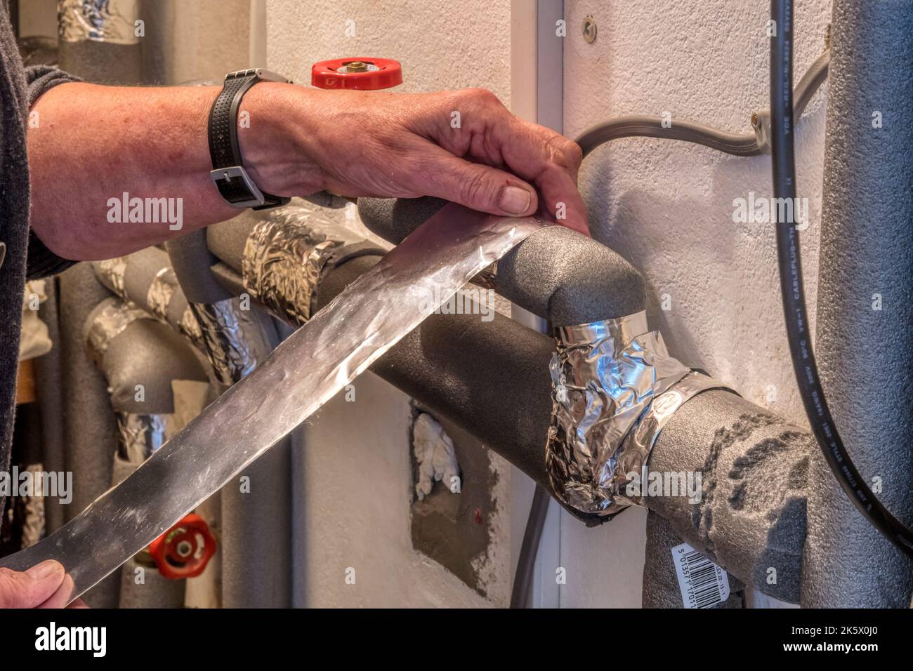 Frau hinkt in einem Belüftungsschrank mit Heißwasserleitungen zurück und klebt sie mit Klebeband, um Wärme zu sparen und die Energieeffizienz zu verbessern. Stockfoto