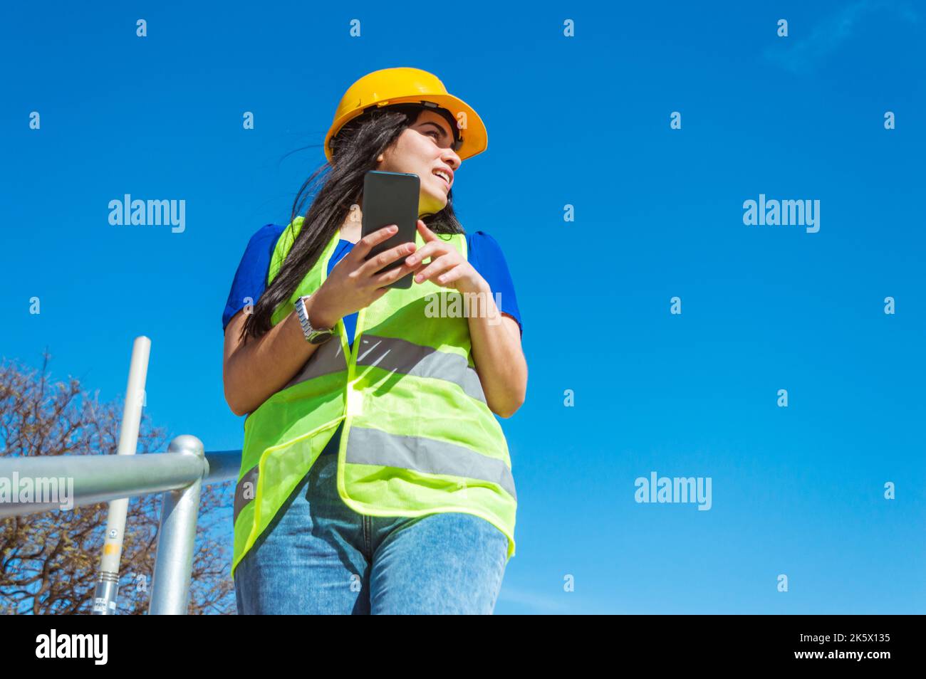 Junge kaukasische Arbeitsfrau mit Schutzhelm steht im Freien und schaut zur Seite, streitet mit jemandem, während sie ihr Telefon hält. Stockfoto