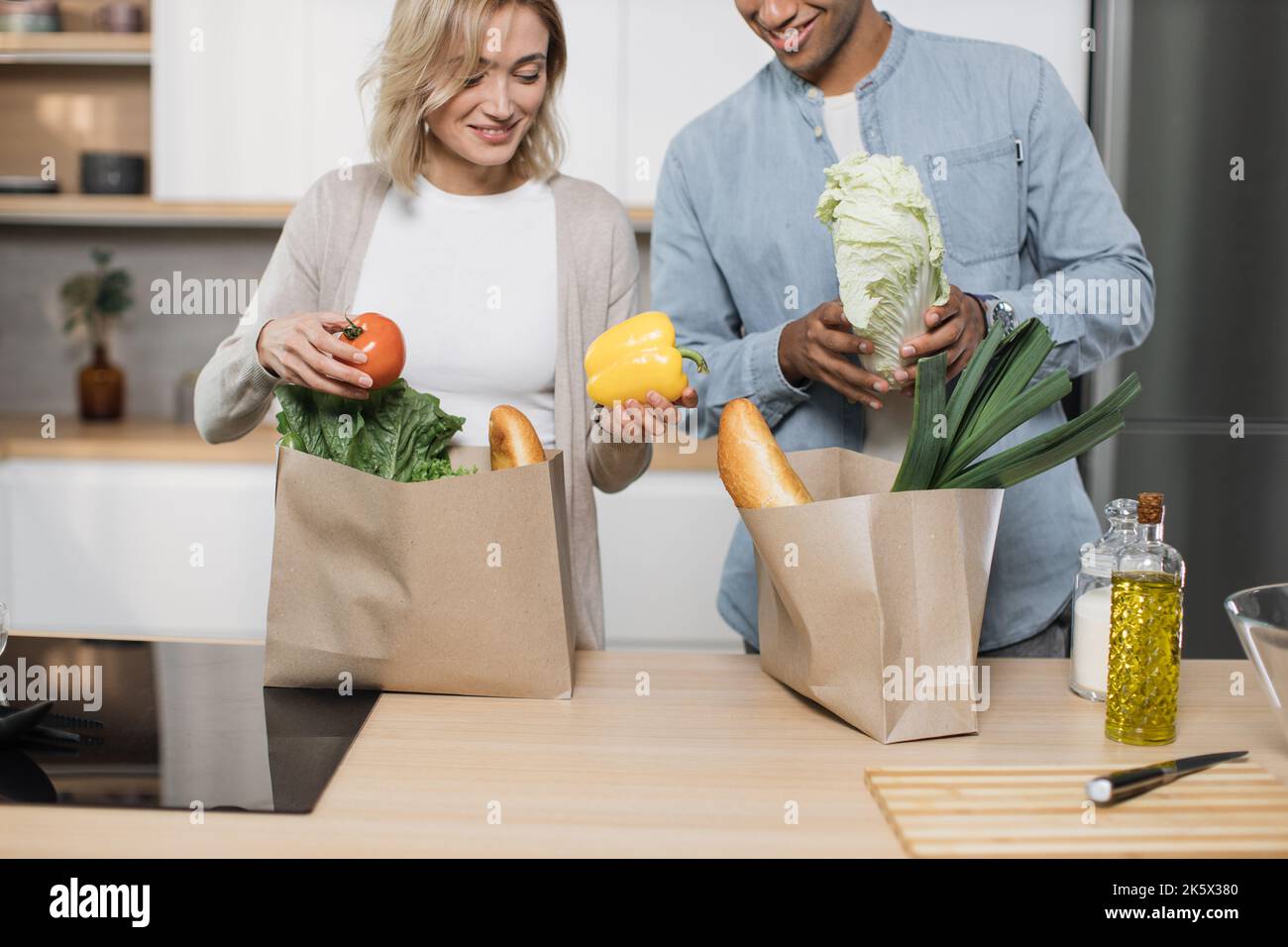 Beschnittenes Bild eines glücklichen jungen Ehepaares, das vom Einkaufen nach Hause kommt und zusammen Papiertüten mit Lebensmitteln in der Küche auspackt. Haushalts- und Beziehungskonzept. Stockfoto