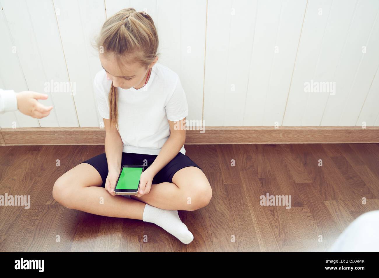 Ein Mädchen, das ein Mobiltelefon mit einem Touchscreen und einer Farbtaste verwendet. Das Kind sitzt auf dem Boden und hält ein Smartphone. Ein unbekanntes Baby zieht seine Hand von der Seite. Hochwertige Fotos Stockfoto