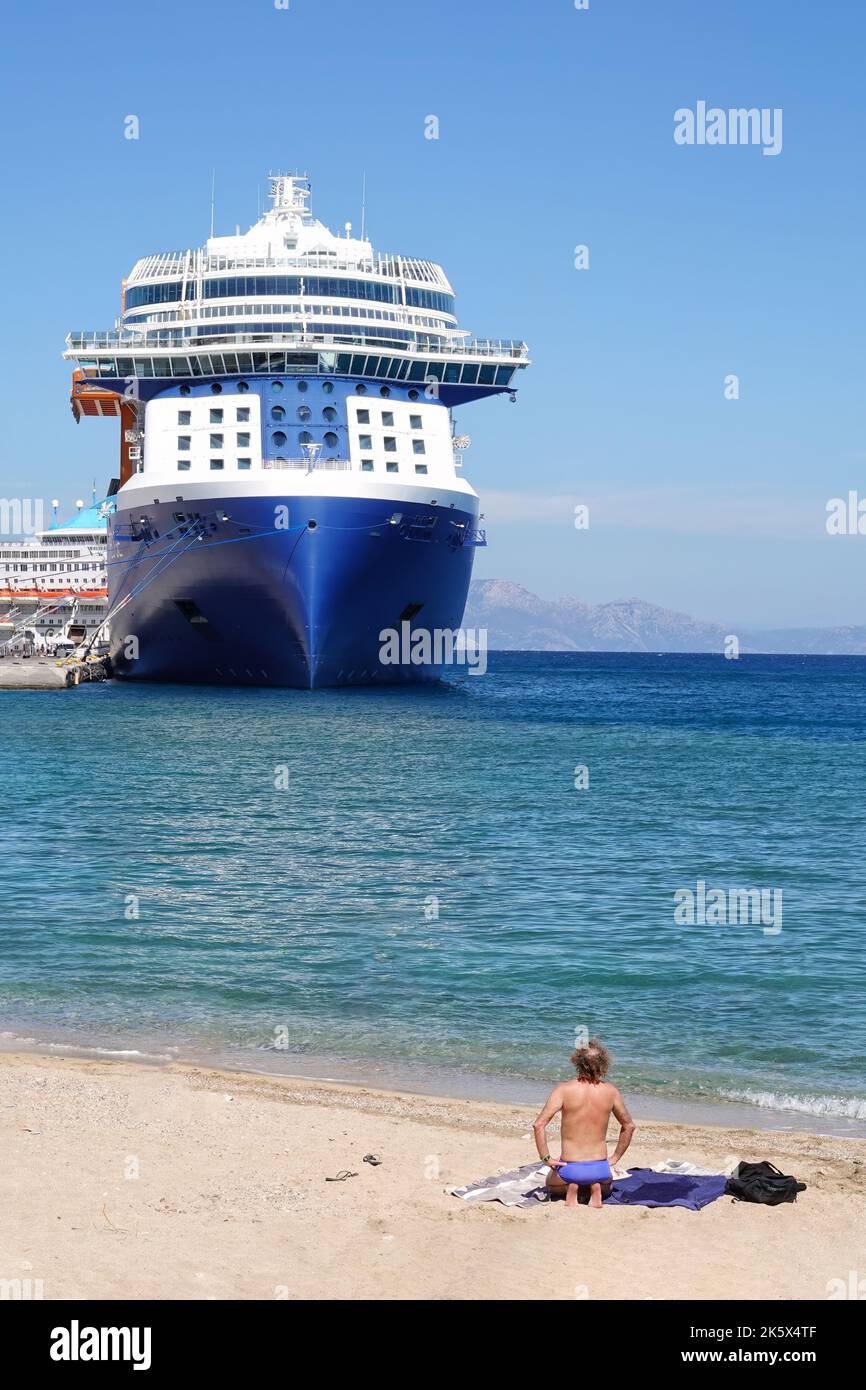 Ein Mann, der an einem Strand sonnenbadet, steht auf den Knien und sieht ein großes Kreuzfahrtschiff, das in der Nähe des Strandes in Rhodos-Stadt, Rhodos, Griechenland, vor Anker liegt Stockfoto