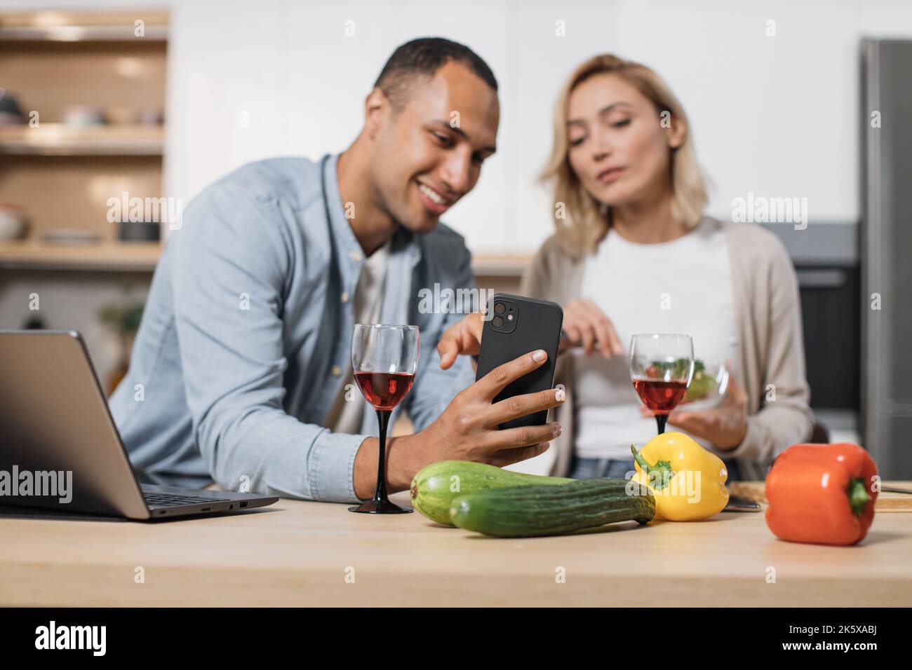 Konzentrieren Sie sich auf das Handy eines glücklichen multinationalen Paares, das Salat isst, und checken Sie Fotos in sozialen Medien, Netzwerken und lächeln Sie in ihrer modernen, hellen Küche zu Hause. Stockfoto