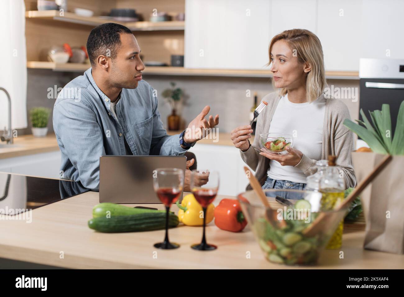Fröhliches multinationales Paar mit Laptop, das Diätsalat aus frischem Bio-Gemüse isst und mit einander spricht, lächelt und entspannt nach dem Arbeitstag in der modernen Küche zu Hause. Stockfoto