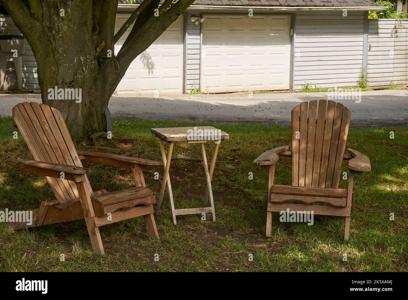 Zwei leere Adirondack-Holzstühle und ein Klapptisch sitzen im Schatten eines Baumes Stockfoto