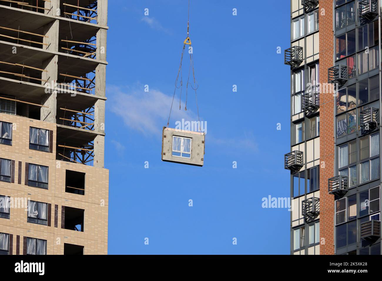 Baukran mit Wand- und Fensterabschnitt zwischen neuen und unfertigen Wohngebäuden auf dem Hintergrund des blauen Himmels. Gehäusekonstruktion Stockfoto