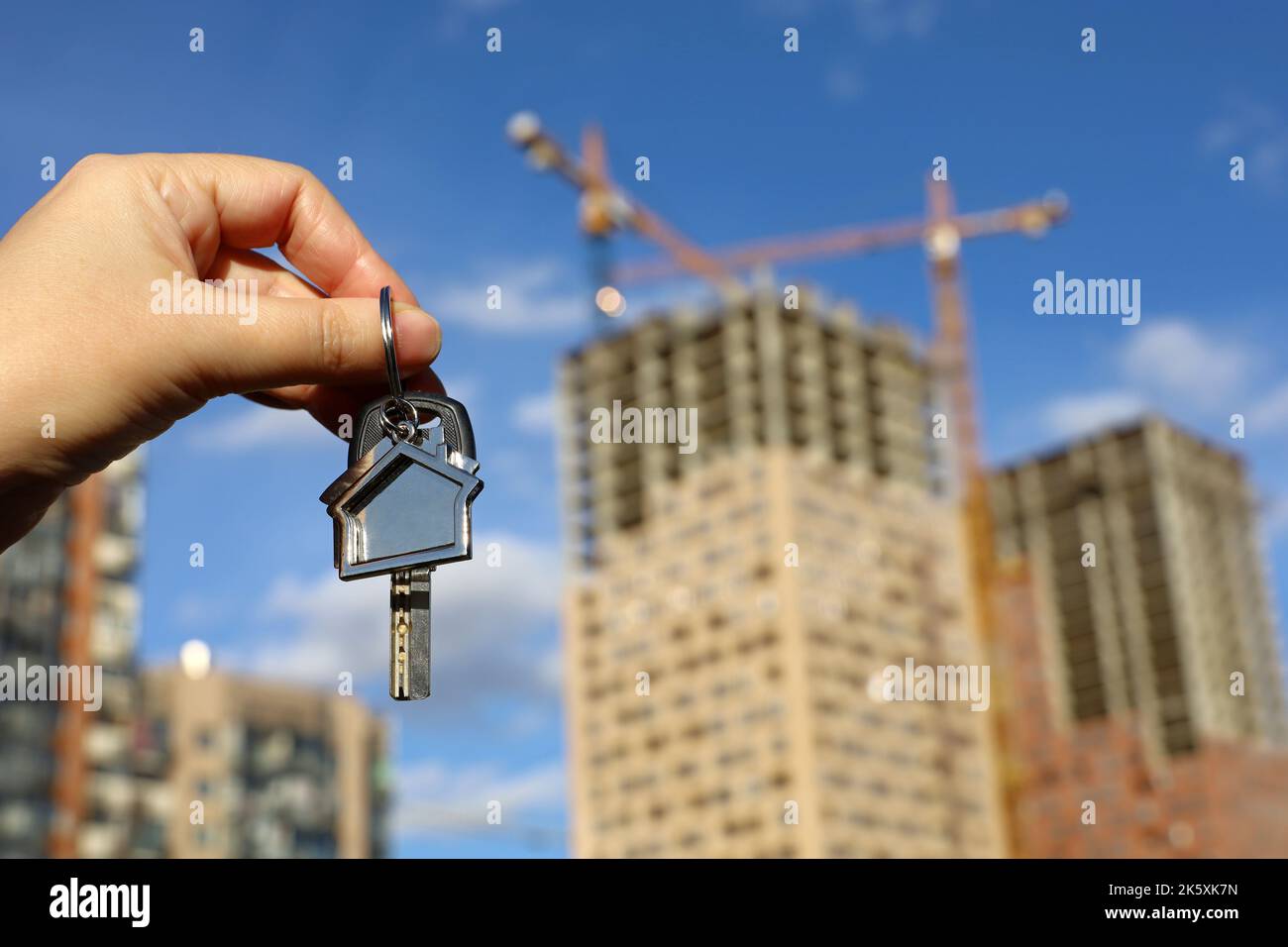 Immobilienmakler, Schlüsselanhänger in Form eines Hauses und Schlüssel in weiblicher Hand auf dem Hintergrund von Baukräne und Neubauten. Byung Wohnung oder Miete Stockfoto