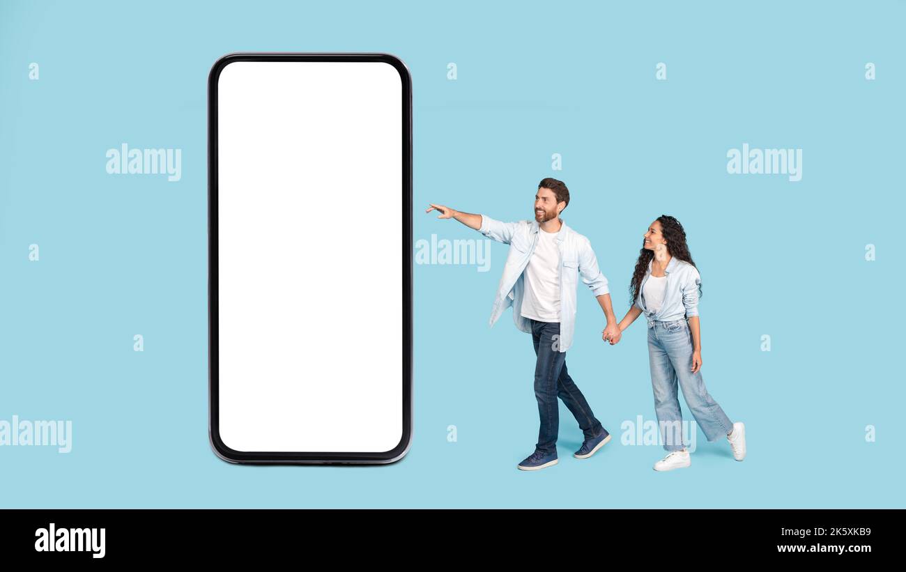 Fröhlicher junger kaukasischer Ehemann hält die Hand der arabischen Frau und zeigt mit dem Finger auf ein riesiges Smartphone mit leerem Raum Stockfoto