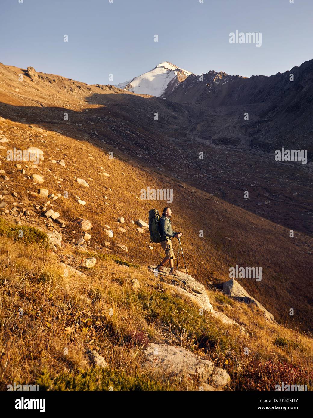 Portrait eines Bartwanderers mit großem Rucksack und Trekkingstöcken, der gegen den wunderschönen Sonnenuntergang im Bergtal den Felsen hinunter läuft. Out Stockfoto