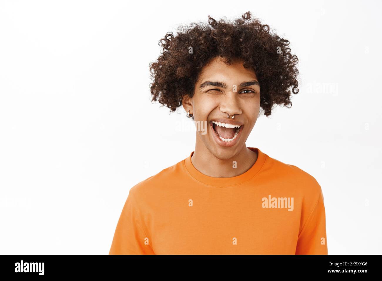 Nahaufnahme eines gutaussehenden hispanischen Mannes mit lockigen Haaren, lächelnd und lachend, frech aussehend, mit orangefarbenem T-Shirt, stehend über weißem Studiohintergrund Stockfoto