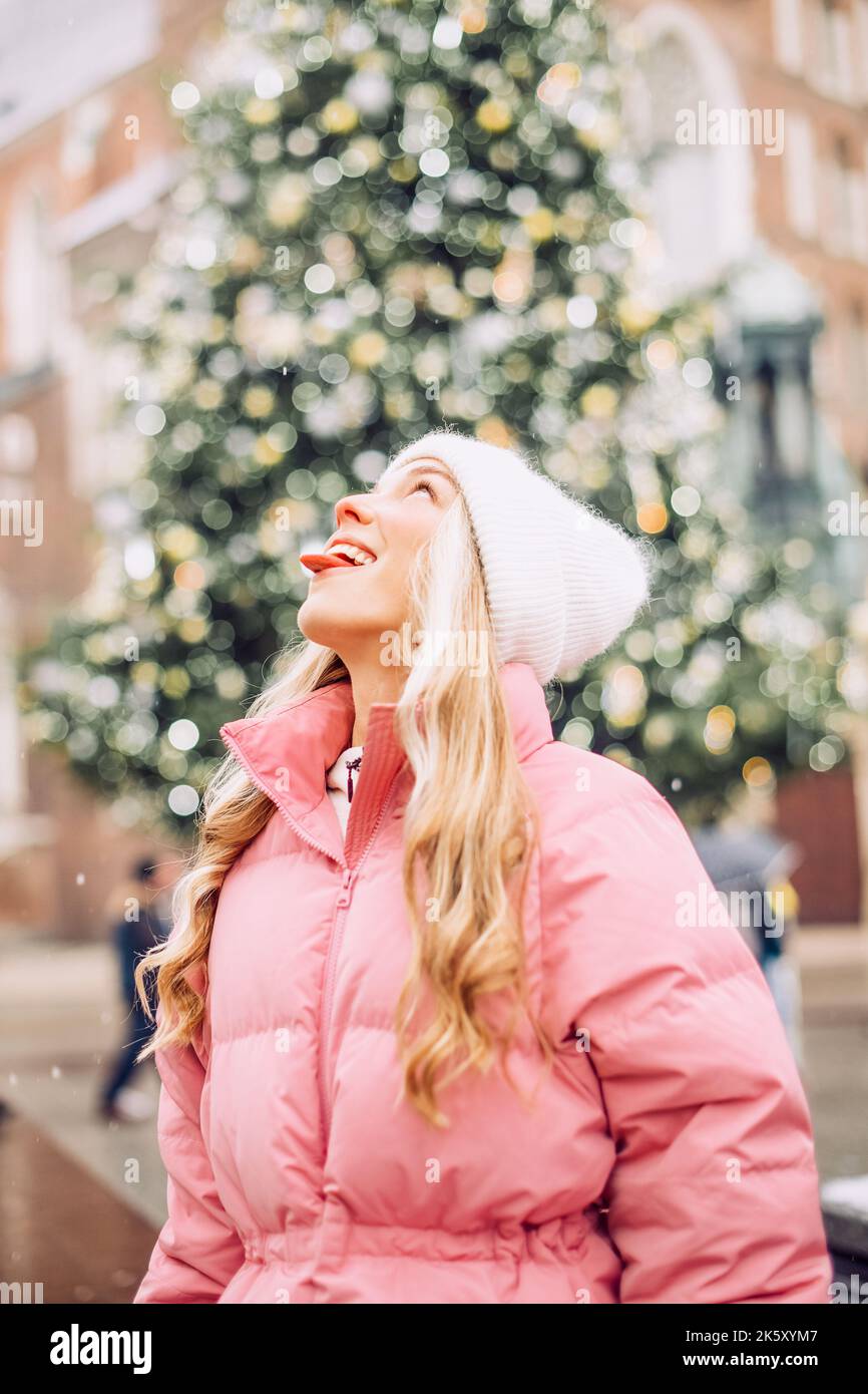 Ein niedliches Porträt eines Mädchens im Winter auf der Straße, der Schnee fällt, die Blondine auf dem Hintergrund des Weihnachtsbaums fängt Schneeflocken mit Stockfoto