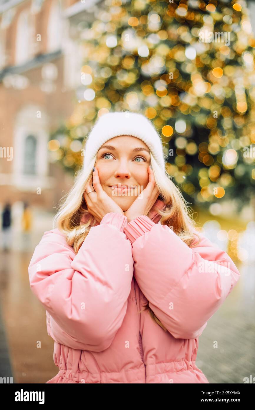 Eine niedliche Blondine in einem Hut und einer rosa Jacke vor dem Hintergrund eines Weihnachtsbaums auf der Straße. Winterspaziergang, Neujahr, Festtag Stockfoto