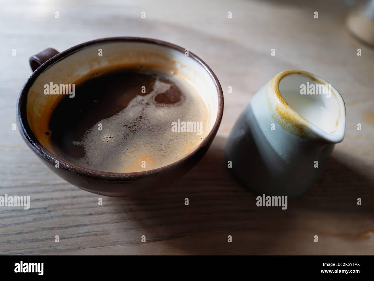 Flacher Fokus auf schwarzen Kaffee in einem rustikalen Stil Töpferbecher neben Milch in einem kleinen Krug. Es ist auf einem bewaldeten Tisch, der durch eine Fensterbeleuchtung mit int beleuchtet wird Stockfoto