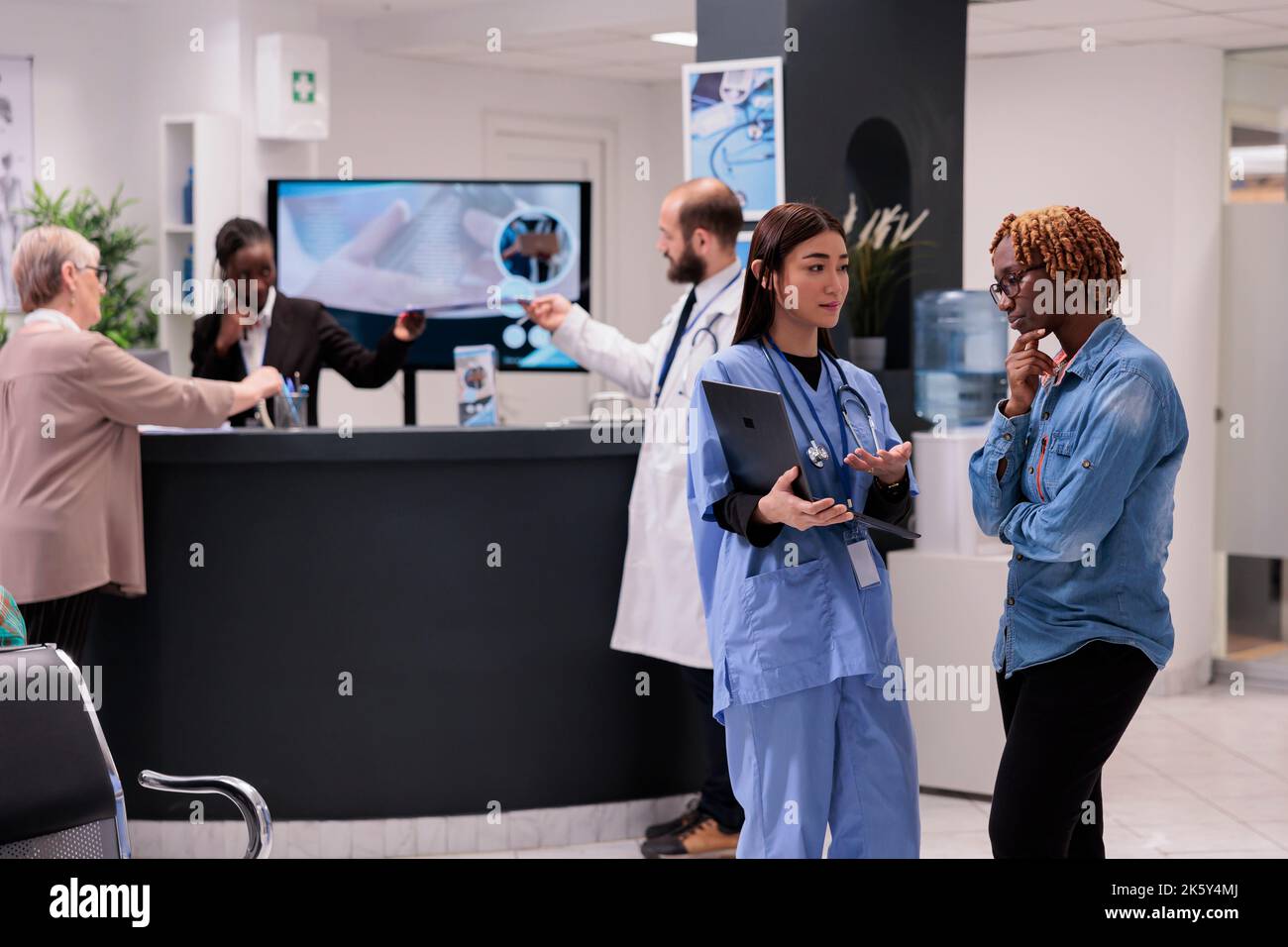 Asiatisches medizinisches Personal teilt klinische Bilder auf dem Laptop mit afroamerikanischen Patienten an der Krankenhausaufnahme. Junge Rezeptionistin, die einen Arzt besucht, ältere Frau am Schreibtisch. Stockfoto