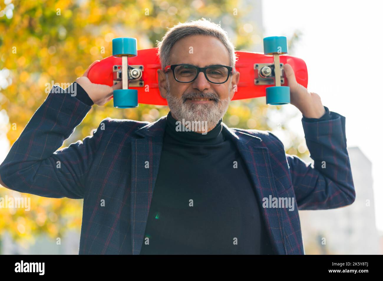 Voller lebenspositiver kaukasischer Großvater mit grauem Gesichtshaar und schwarzer Brille in dunklem Blazer und Rollkragen, der die Kamera anschaut und ein leuchtendes orangefarbenes kleines Skateboard seines Enkelkindes mit zwei Händen hinter dem Kopf hält. Außenaufnahme. Aktiver Herbst. Hochwertige Fotos Stockfoto