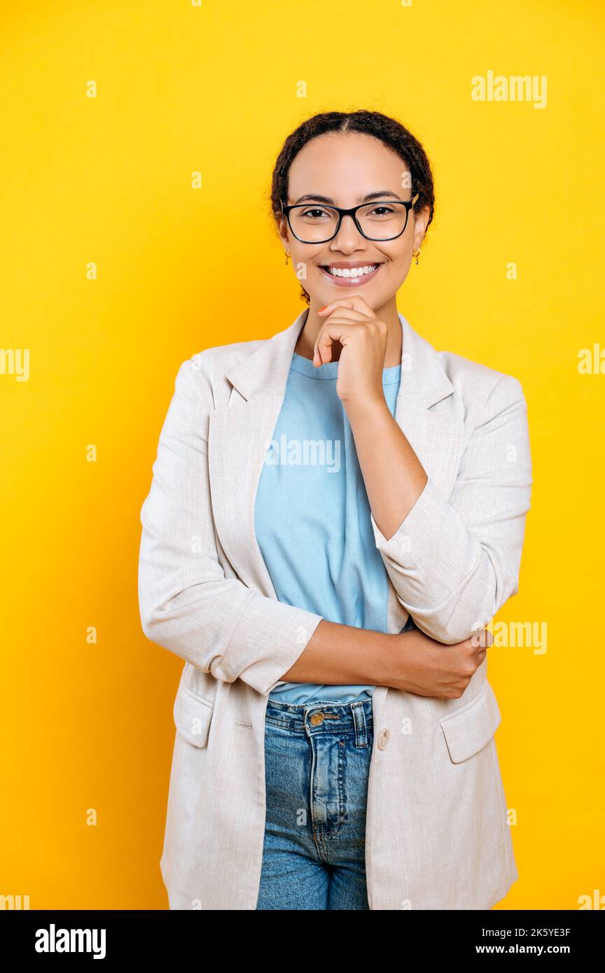 Vertikales Porträt einer ziemlich erfolgreichen hispanischen oder brasilianischen Frau mit Brille, stilvoll gekleidet, hr-Manager, Immobilienmakler, Makler, Blickt auf die Kamera, lächelt, steht auf einem isolierten orangefarbenen Hintergrund Stockfoto