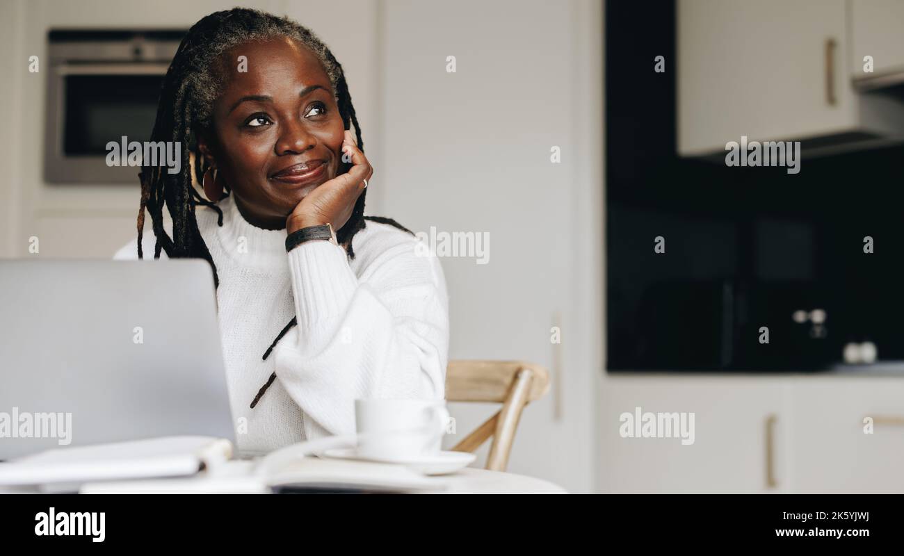 Eine reife Geschäftsfrau blickt bei der Arbeit an einem Laptop zu Hause nachdenklich weg. Leitende Geschäftsfrau, die freiberuflich im Internet in ihrem Heimbüro arbeitet. Stockfoto