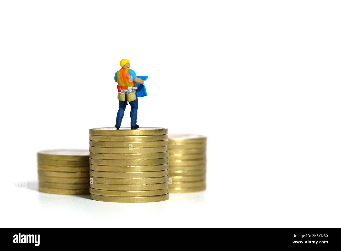 Miniatur Menschen Spielzeug Figur Fotografie. Projektbudgetkonzept. Ein Bauarbeiter mit einem Blaupause-Gebäude, das über dem Goldmünzenstapel steht. Stockfoto