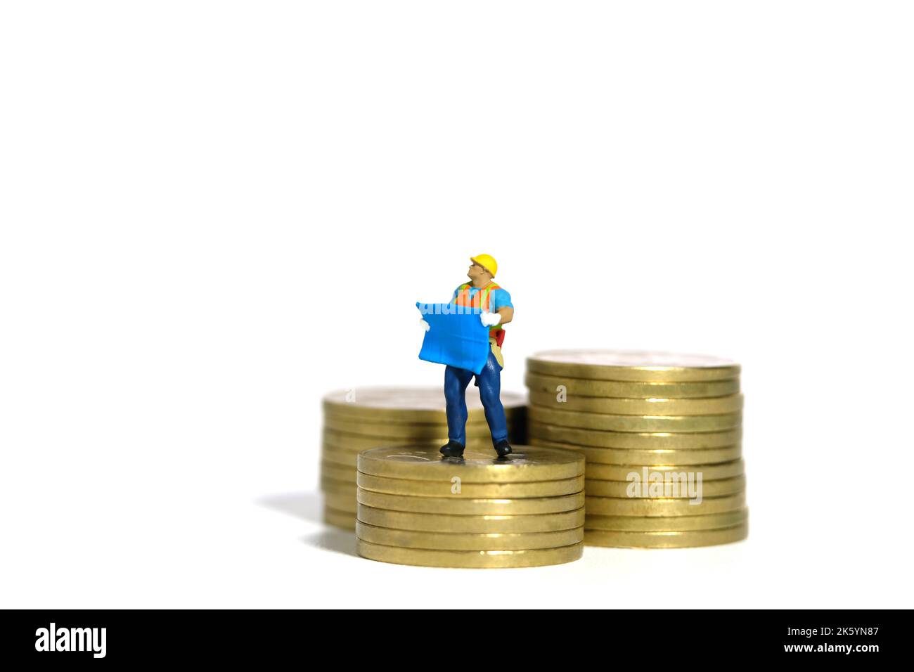 Miniatur Menschen Spielzeug Figur Fotografie. Projektbudgetkonzept. Ein Bauarbeiter mit einem Blaupause-Gebäude, das über dem Goldmünzenstapel steht. Stockfoto