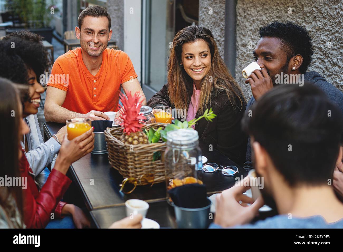 Eine Gruppe junger, multiethnischer junger Leute beim Frühstück, die sich alle unterhalten und beim Brunch-Frühstück Spaß haben - verschiedene Leute im Café, die Obst trinken j Stockfoto