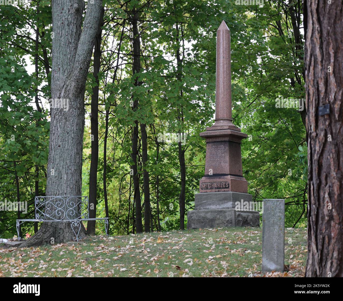 Ein großer Obelisk und ein kleiner Grabstein sitzen auf einem Hügel, umgeben von großen Bäumen. Der Herbst hinterlässt auf dem Boden. Ruhig und besinnlich Stockfoto