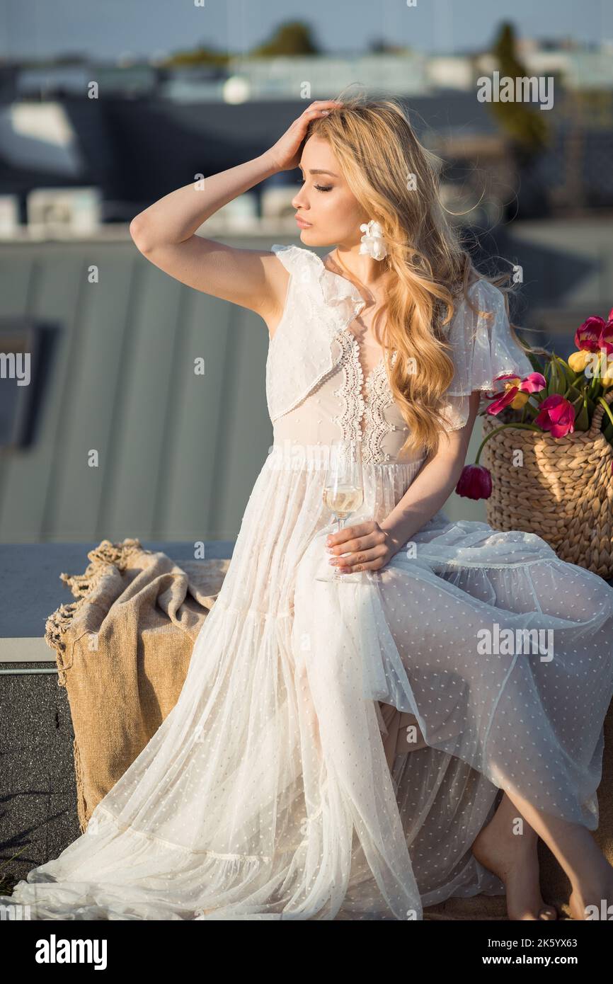 Attraktive Frau in stilvollem weißen Kleid sitzt auf einer Decke und mit gedrehtem Kopf Blick auf den Sonnenuntergang auf der Dachterrasse. Elegante Dame entspannen bei einem Glas Champagner Stockfoto