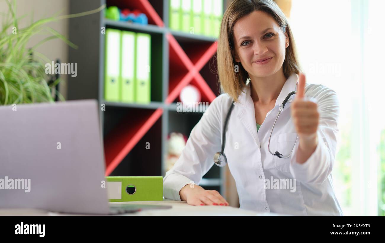 Fröhliche Ärztin posiert im Klinikbüro und zeigt mit Daumen nach oben Zeichen Stockfoto
