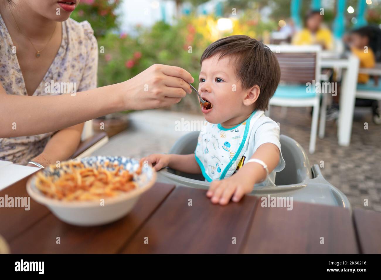 Gutaussehender Mischling ein Jahr alter Junge, der ein Abendessen im Restaurant mit seiner Mutter, die ihn in einem nicht erkennbaren Restaurant am Abend füttert, zu sich nahm Stockfoto