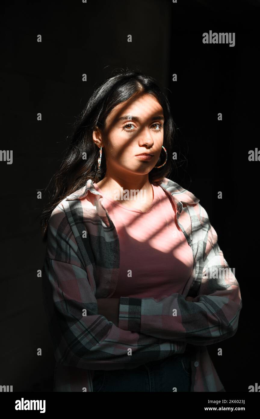 Selbstbewusste junge Frau in legerer Kleidung, die die Arme kreuzt und die Kamera anschaut, während sie im dunklen Raum unter Sonnenlicht vor schwarzem Hintergrund steht Stockfoto