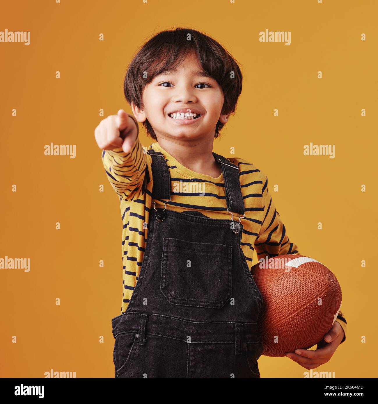 Netter kleiner gemischter Rennjunge, der lächelt und zeigt, während er einen Fußball hält und vor einem orangen Copyspace-Hintergrund posiert Stockfoto