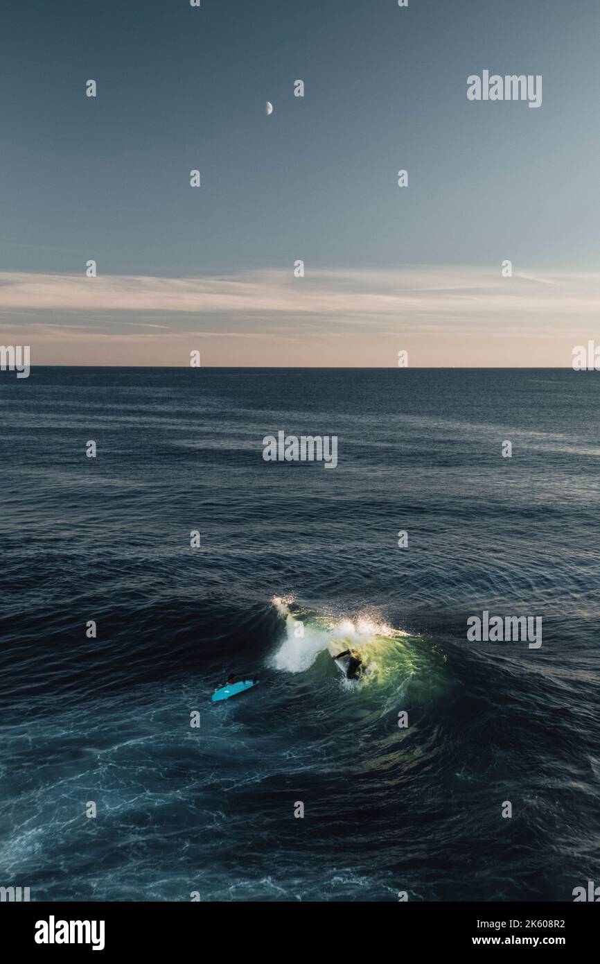 Abenteuerlustige Surfer, die eine Welle unter dem Abendhimmel mit einem Mond und weit entfernten Klippen in Cornwall einfangen, um das Wesen des Surfens einzufangen. Stockfoto Abenteuerlustige Surfer, die eine Welle unter dem Abendhimmel mit einem Mond und weit entfernten Klippen in Cornwall einfangen, um das Wesen des Surfens einzufangen. Stockfoto