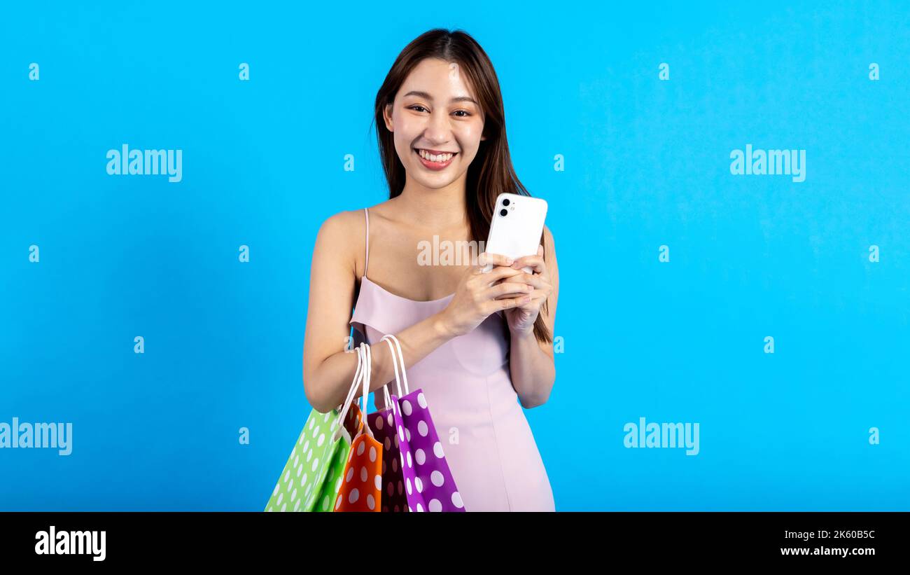 Glücklich lächelnde asiatische weibliche Shopper hält eine Reihe von Einkaufstaschen, während ein Mobiltelefon hält. Modische Mädchen in Kleid Blick auf Kamera isoliert auf blauem Hintergrund - mit Kopierraum Stockfoto