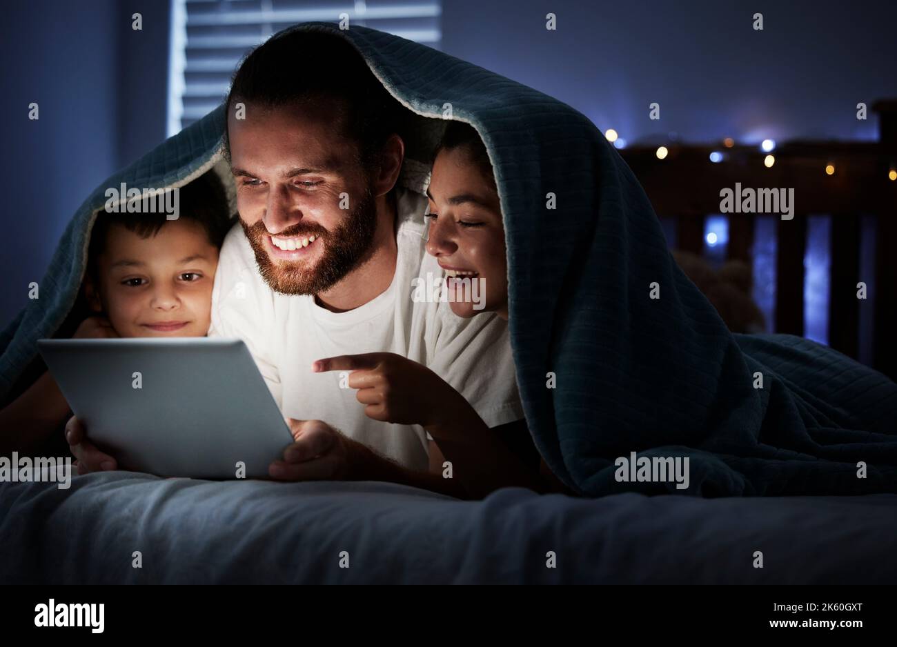 Glücklicher kaukasischer Familienvater mit zwei Kindern, die ein digitales Tablet benutzen, das in der Nacht unter einer Decke im Dunkeln liegt und deren Gesichter von ihnen erleuchtet sind Stockfoto
