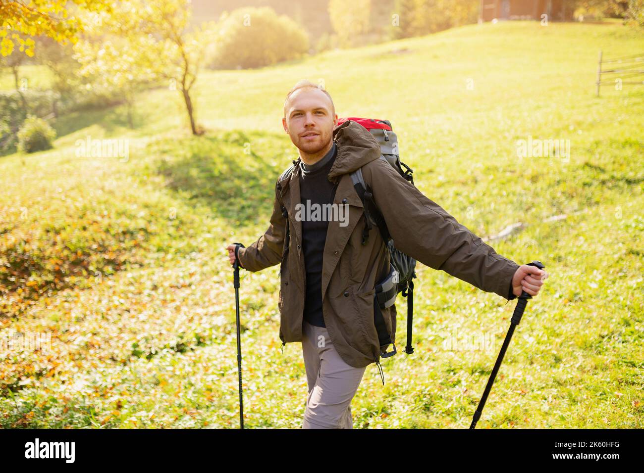 Wandermännchen mit Wanderausrüstung steht mitten in einem Bergdorf auf einem Hügel und blickt auf die Kamera. Glücklicher Kerl mit Rucksack und Trekking Stockfoto