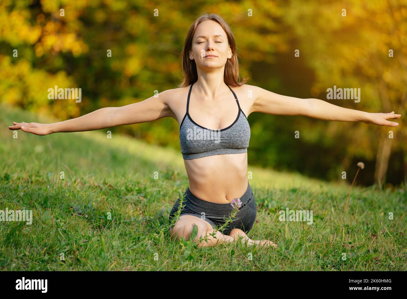 Ruhige, schlanke junge Frau, die mit geschlossenen Augen in einer Yoga-Pose mit ausgestreckten Armen auf dem Gras sitzt. Konzept für Alltag, Fitness, y Stockfoto