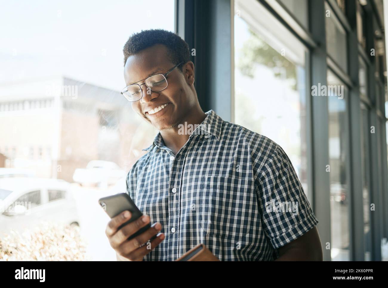Glücklicher afroamerikanischer Geschäftsmann, der sich in einem Büro an ein Fenster lehnt, während er Text auf dem Smartphone liest oder sendet. Lächelnd Stockfoto