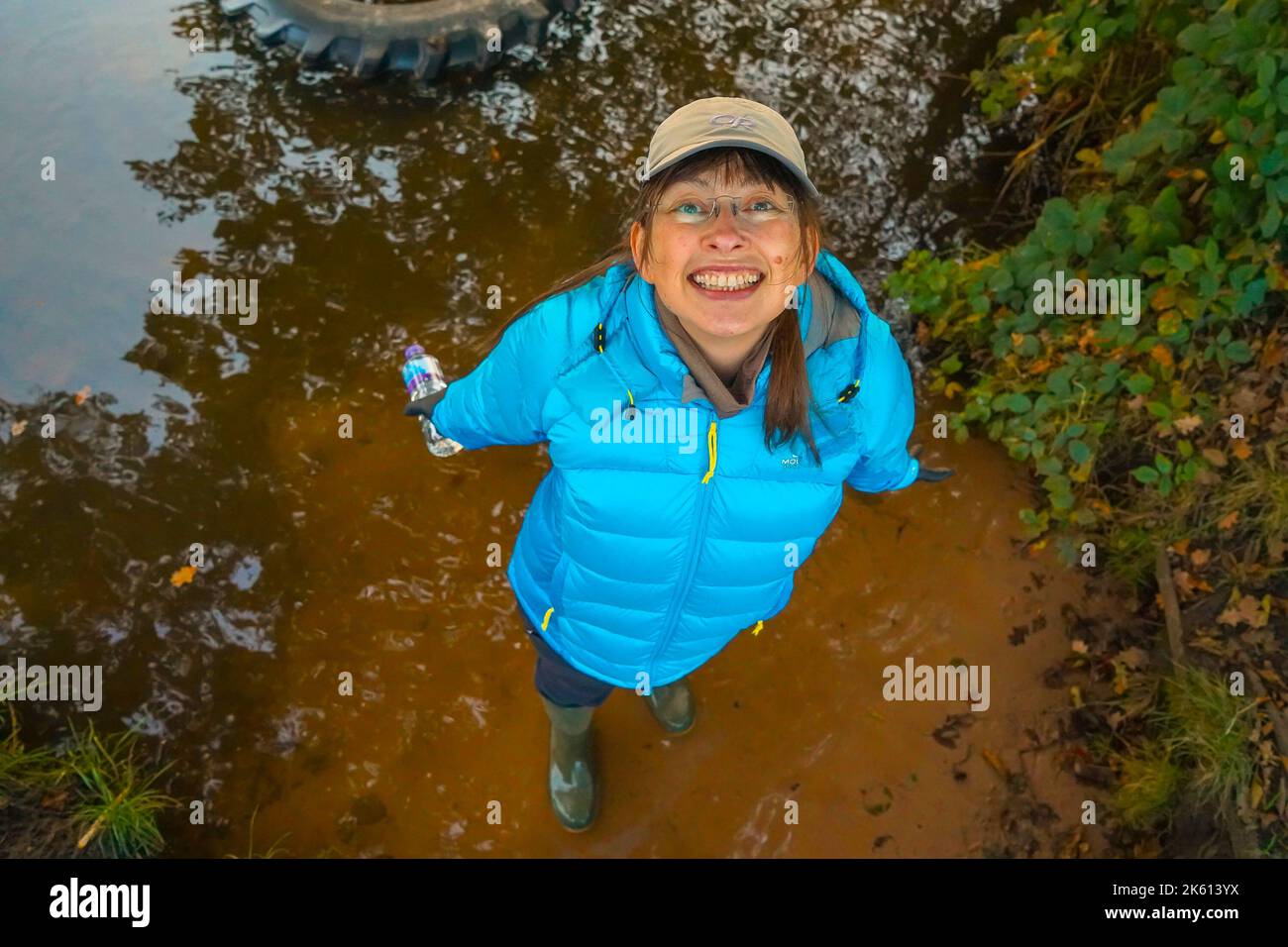 Blick von oben auf eine lächelnde Frau in Gummistiefel, die nach oben schaut und in Regenwasserpfützen steht. Stockfoto