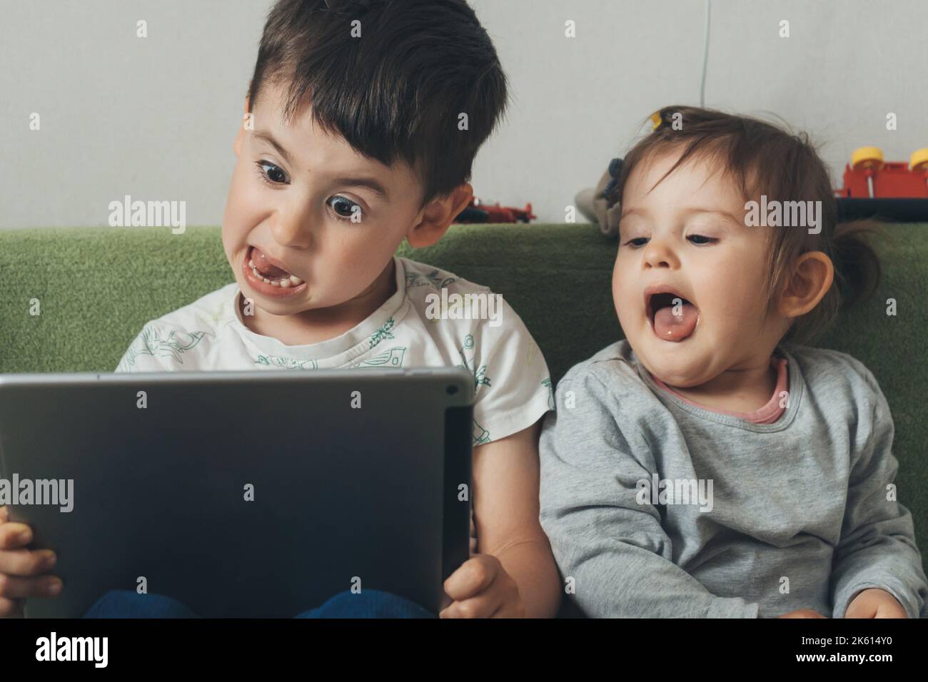 Junge und Mädchen spielen auf einem Tablet, während sie in ihrem Zimmer auf dem Sofa sitzen und unterschiedliche Gesichtsausdrücke machen. Technologiekonzept. Glückliches Familienkind Stockfoto